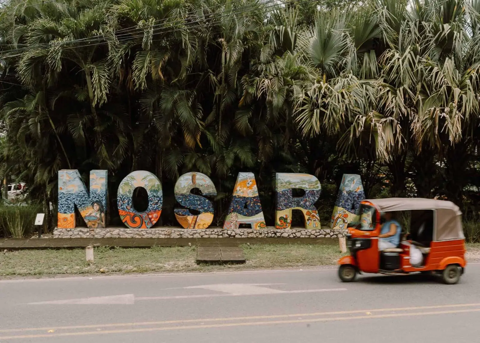 The painted NOSARA letters near the main road, tuk tuk rolling past